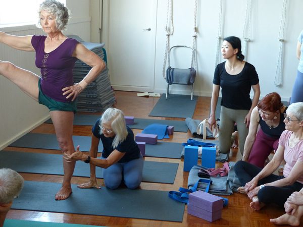 Pixie instructing a group of students. One student is demonstrating utthita hasta padangustasana, Pixie is pointing at their knee